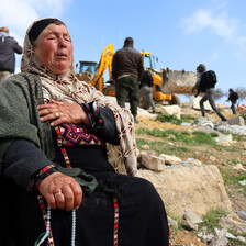 Woman sits on a rock with her hand on her chest as bulldozer operates in the background 