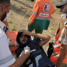 A wounded man wearing a press helmet and flak jacket is carried on a stretcher