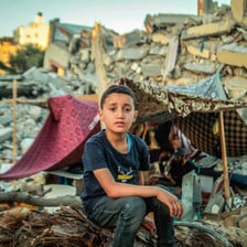 Boy sits on rubble in front of makeshift tent amid destroyed buildings