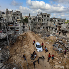 Aerial view of a car and people on a dirt-covered road surrounded by bombed-out buildings