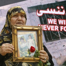 Woman holds framed photo of young man and rose in front of banner reading We will never forget in English and Arabic