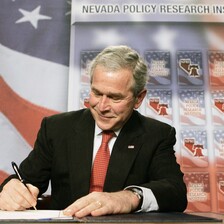 George W. Bush smirks as he signs document while sitting at table with American flag backdrop behind him