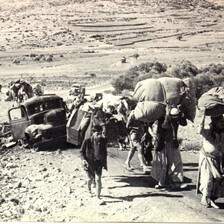 A black and white photo shows people on foot carrying heavy burdens passing a broken down vehicle