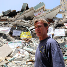 Man wearing T-shirt stands in front of destroyed building 
