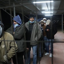 Men, some wearing masks, line up behind a grille 
