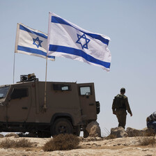 Two blue and white flags fly beside an armored vehicle and soldiers in uniform 