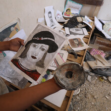 A portrait of a woman wearing a hat is held by a hand while some other pictures are laid out on a wooden bed