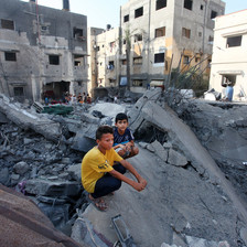Two boys sit on ruins of fallen building while surrounded by bombed-out midrise buildings