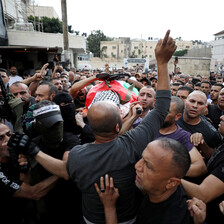 A flag drapes a body on a stretcher carried by a large crowd