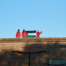 Three people in red hold up a Palestinian flag on a rooftop
