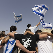 Young men seen with their backs to camera embrace with Israeli flags waving above them. One is wearing a black T-shirt showing the Star of David with a rifle in the middle.