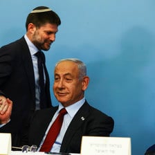 Bezalel Smotrich grasps a seated Benjamin Netanyahu's hand while they both smirk
