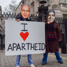 Two people wearing masks mimicking Benjamin Netanyahu and Rishi Sunak hold a sign reading I heart apartheid 