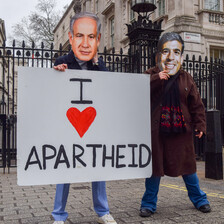 Two people wearing masks mimicking Benjamin Netanyahu and Rishi Sunak hold a sign reading I heart apartheid