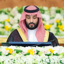 Bin Salman, wearing a head dress and a gold-trimmed cloak, looks down towards a podium while surrounded by flowers