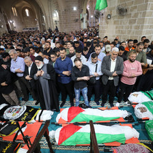 Men stand while praying while facing bodies wrapped in Palestine and faction flags on ground in front of them inside mosque