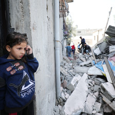 Child wearing a navy sweatshirt stands in the doorway of a building beside a pile of rubble 