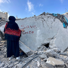 A woman stands amid rubble of a home with the words Save Gaza written in red on a piece of concrete