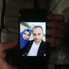 Hands hold a mobile phone showing a picture of a woman wearing a blue scarf and a man a black jacket and a white T-shirt 