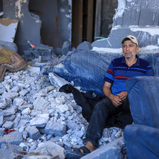 Man wearing a baseball cap sits amid the rubble of a destroyed building 