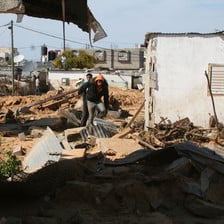 Two young men walk beside a building with much debris on the ground 