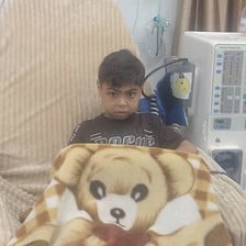 A boy wrapped in a blanket depicting a large teddy bear sits on a chair beside a dialysis machine