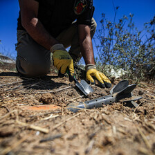 A close-up photograph of an EOD team member with a small shovel digging around a piece of ordnance.