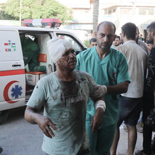 A wounded man with his head wrapped in a bandages is brought into hospital by a medic wearing green overalls
