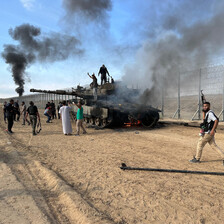 People climb on top of a burning Merkava tank as black smoke and flames rise from it