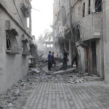 Four men stand along a small road between destroyed homes covered in dust