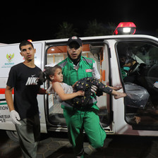 A medic wearing a baseball cap and green overalls carries a girl in front of an ambulance 