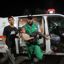 A medic wearing a baseball cap and green overalls carries a girl in front of an ambulance