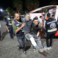 An injured man is carried by two others with an ambulance and medics in the background 