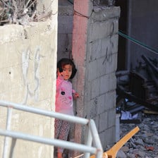 A child wearing a pink bunny sweater peeks out from behind a cinderblock wall surrounded by rubble