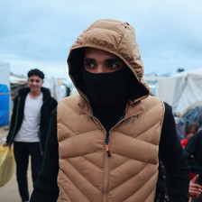 A closeup of a young man wearing a tan puffy hoodie with a mask over his face due to the cold