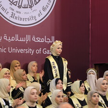 A young woman stands during a graduation ceremony in front of a banner featuring the logo of the Islamic University of Gaza 
