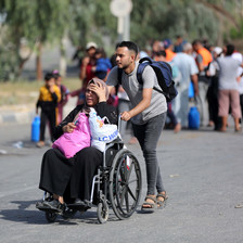 A man pushes a woman in a wheelchair with a group of people on foot behind them