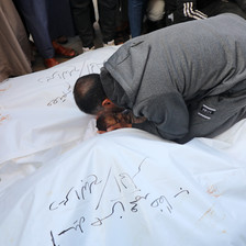 A man kneels down surrounded by dead bodies, most covered by white shrouds 