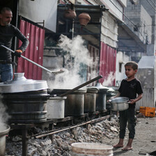 A child waits with a bowl in front of pots of food cook over large wood-burning fires 