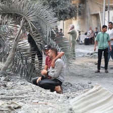 Two men kneel beneath a destroyed palm tree, covered in soot