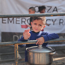 A boy with a pot in front of a banner with the words Gaza Emergency 