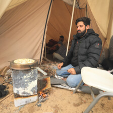 Outside of a tent, a man cooks bread on an outdoor stove