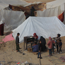 A family tries to heat up some food on a stove beside a number of tents 
