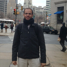 Refaat seen from waist up stands in front of a city crosswalk while wearing a black jacket and looking at the camera