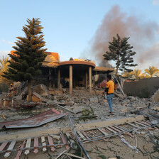 A man stands in front of a destroyed building, smoke rising from behind