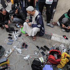A group of men sit on the street around a power cord, charging phones