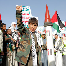 A boy wearing a camouflage jacket and a hatta holds his right fist in the air while standing in a crowd outside