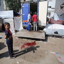 A young boy stands next to a blood stain on pavement and a truck being unloaded.