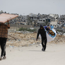 Two people carry some of their belongings on a road with the remains of homes that have been attacked in the background