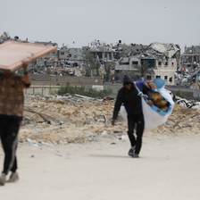 Two people carry some of their belongings on a road with the remains of homes that have been attacked in the background 